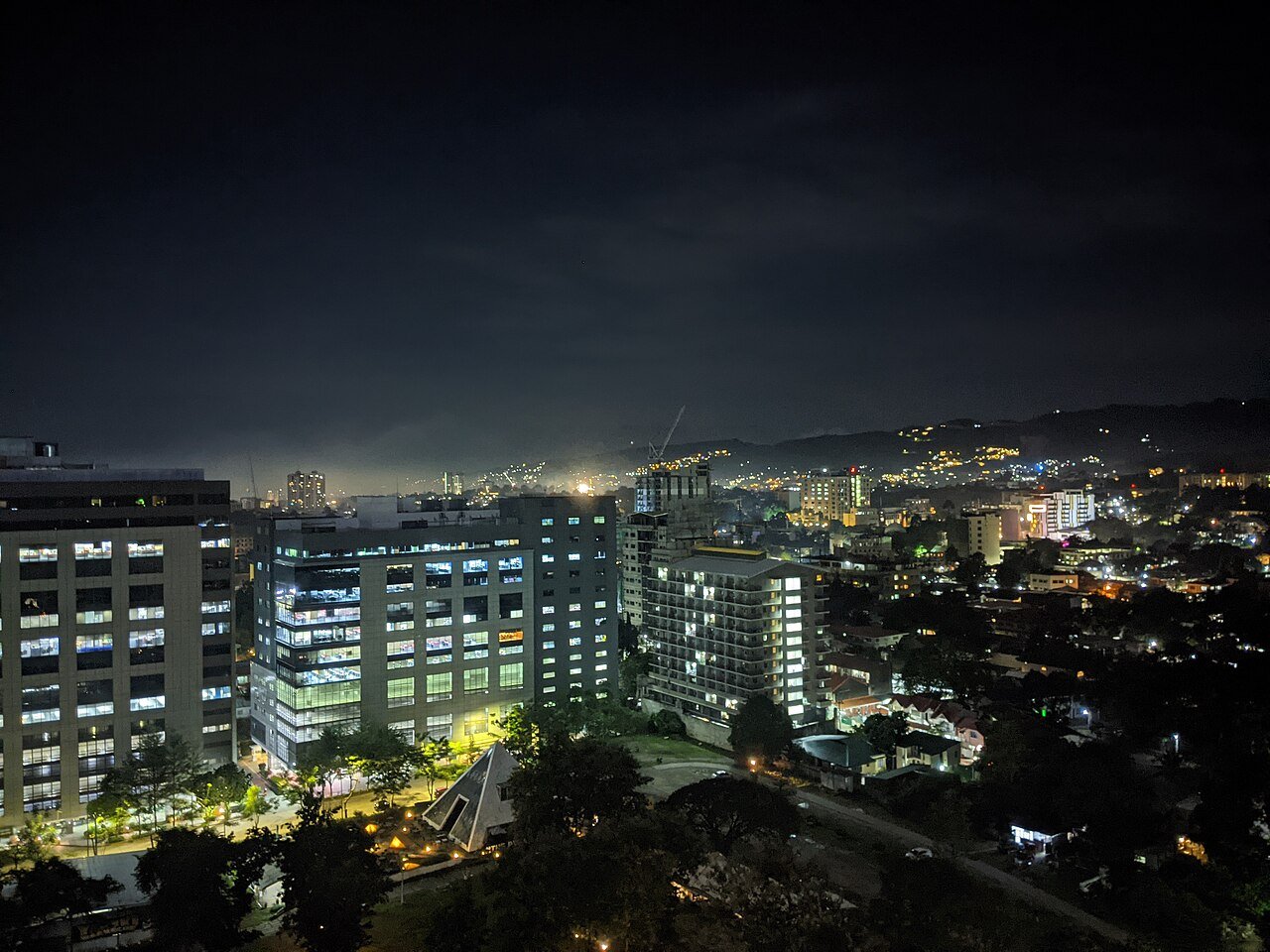 Office towers and a garden with a pyramid-shaped pavilion at night, with hillside lights in the distance