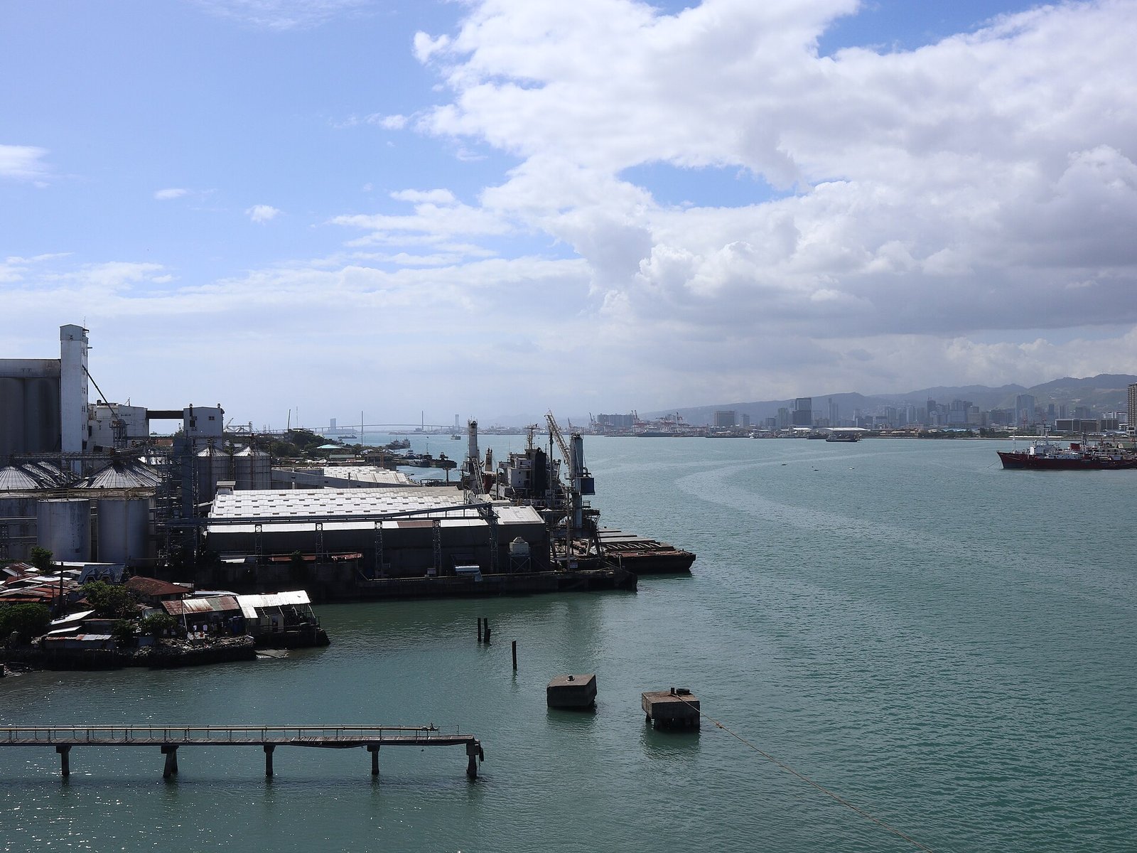 View along a channel past port warehouses and grain silos toward the Cebu City skyline, with a cable-stayed bridge in the distance